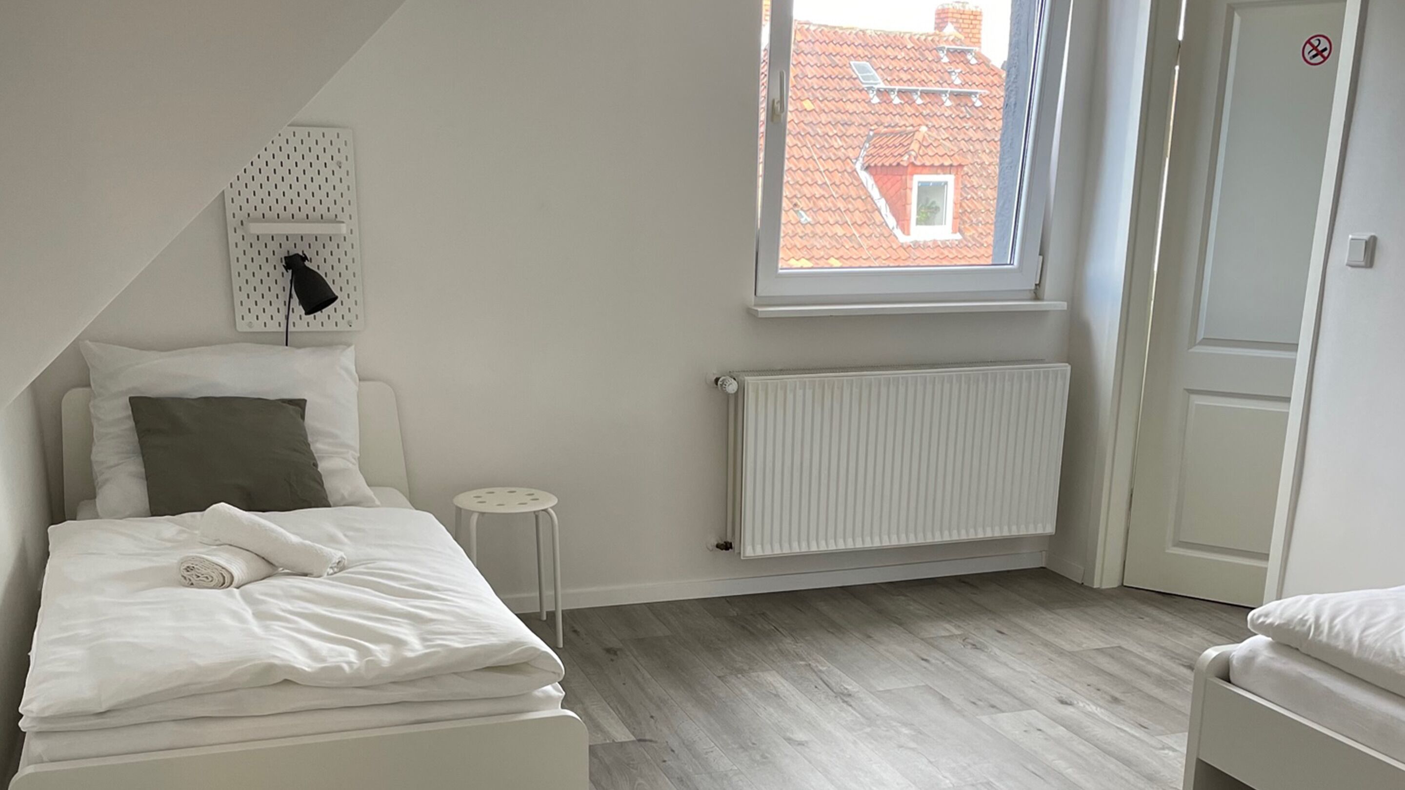 A bedroom with a bed, a radiator, a window with a view of buildings, and a door with a no-smoking sign.