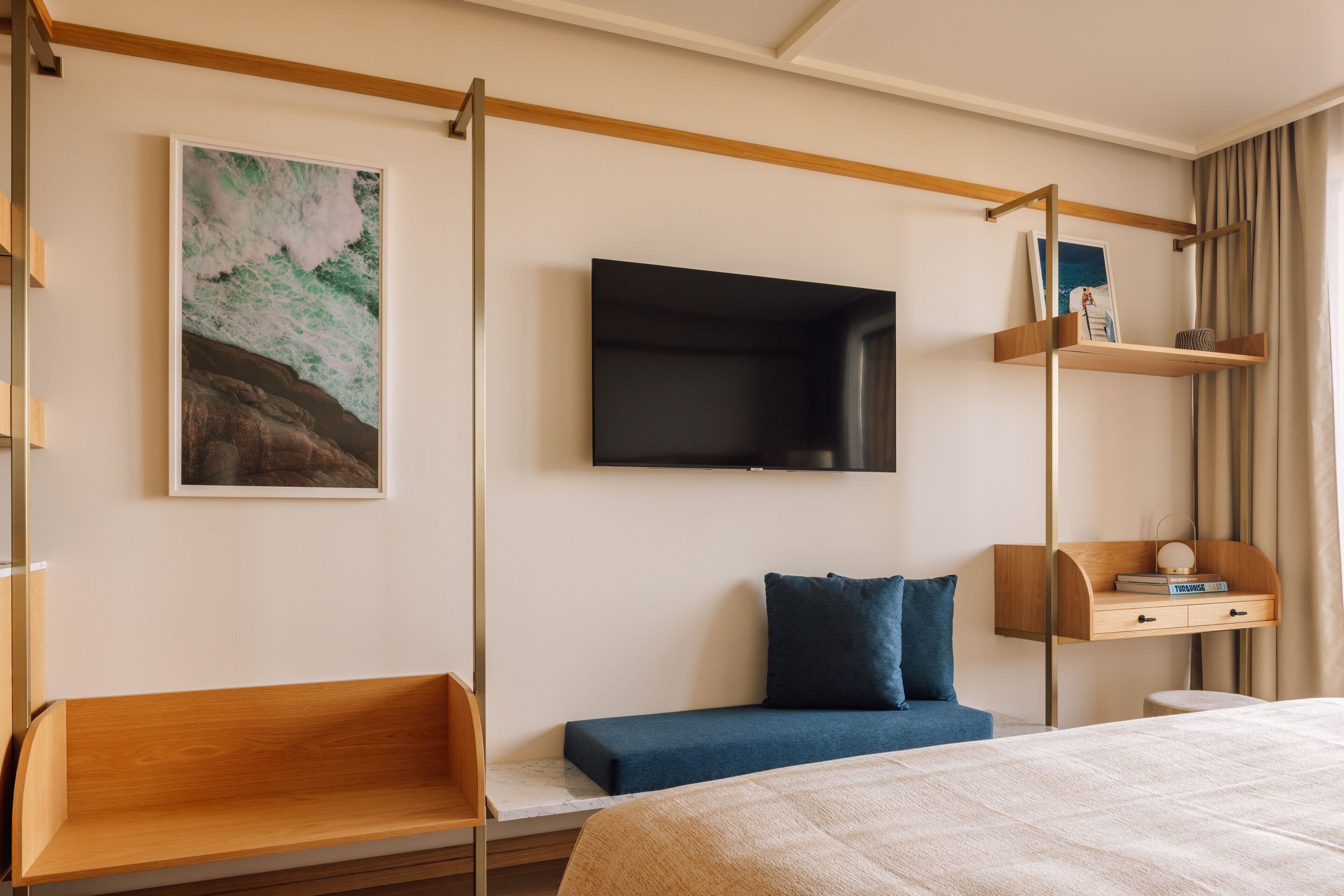 A modern hotel room with a bed, a bench, a TV mounted on the wall, and a shelf with books and a lamp.