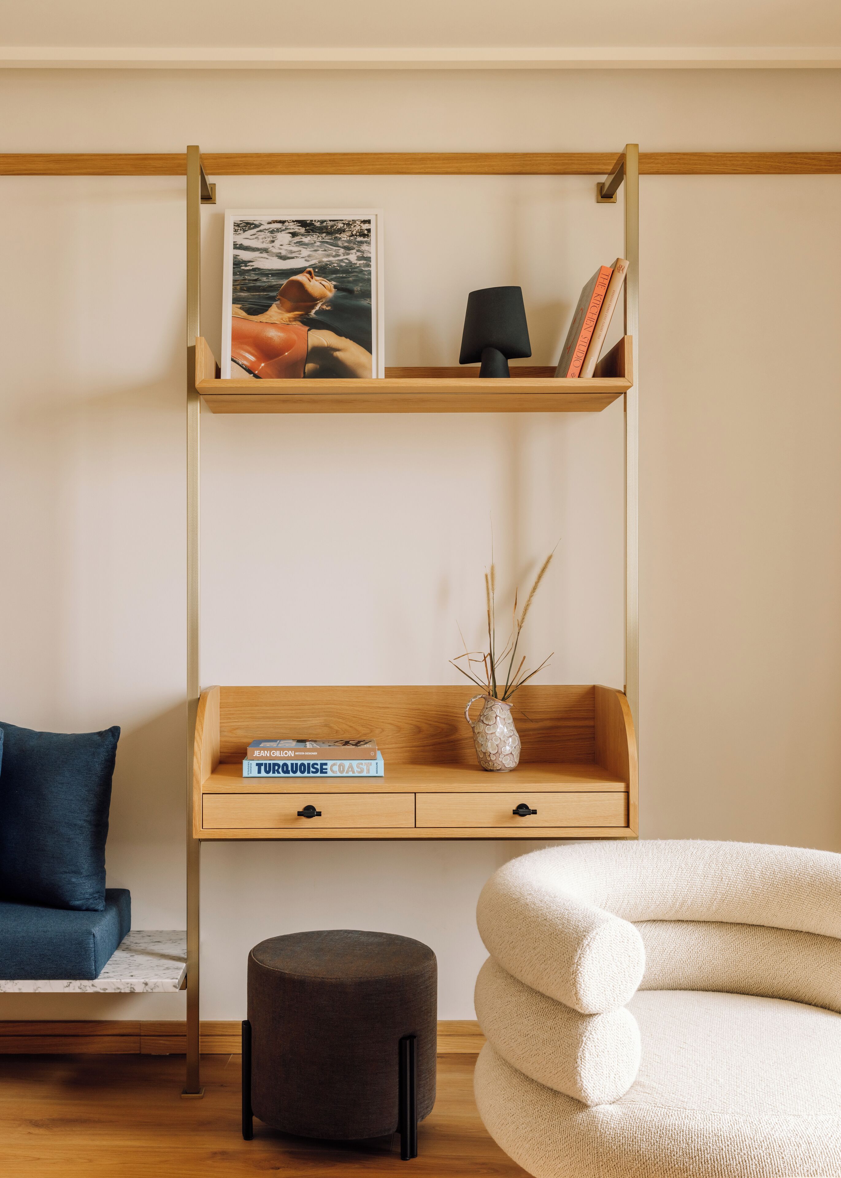 Une chambre moderne avec une étagère en bois, une petite table sur laquelle se trouvent un vase, un livre et un pouf foncé.