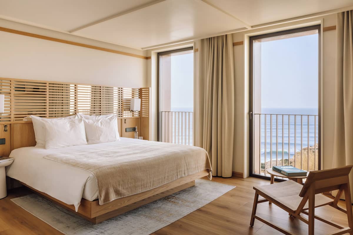 Ein geräumiges Schlafzimmer mit einem großen Bett, einem hölzernen Kopfteil, einem Nachttisch, einem Beistelltisch, einem Sessel und Meerblick durch eine Schiebetür.