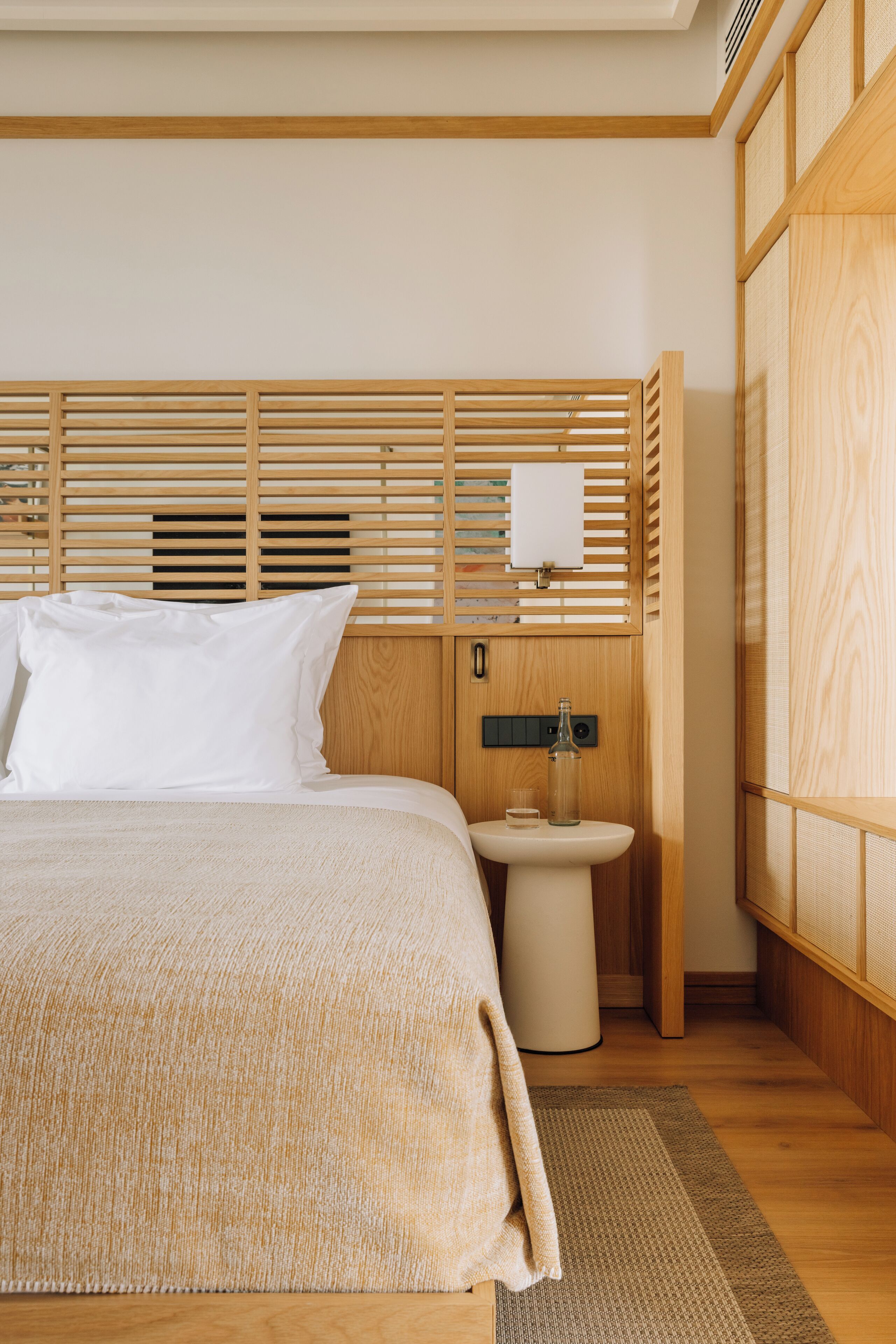 A neatly arranged bedroom with a wooden headboard, a bedside table with a bottle and a candle, and a small sink.