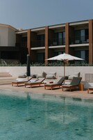Zona de piscina con sillones de descanso y sombrillas, una piscina cristalina y un edificio con varios balcones al fondo.