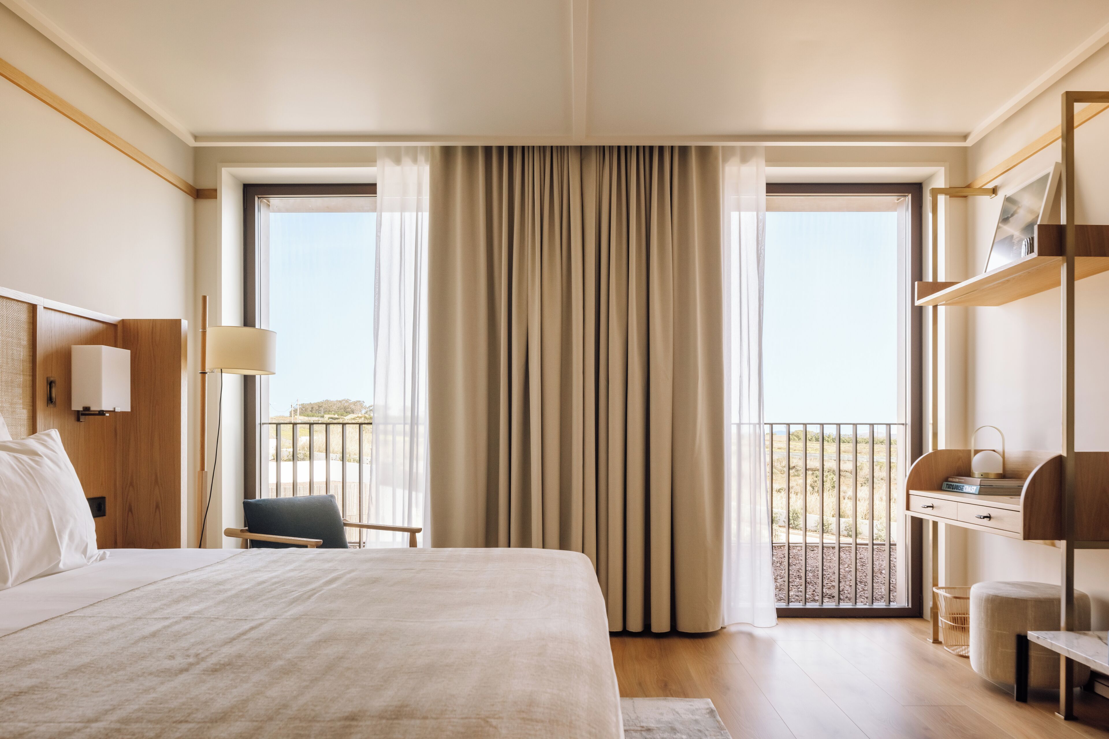 Habitación de hotel con una cama grande, un escritorio con lámpara, una silla y vistas al exterior desde un balcón.