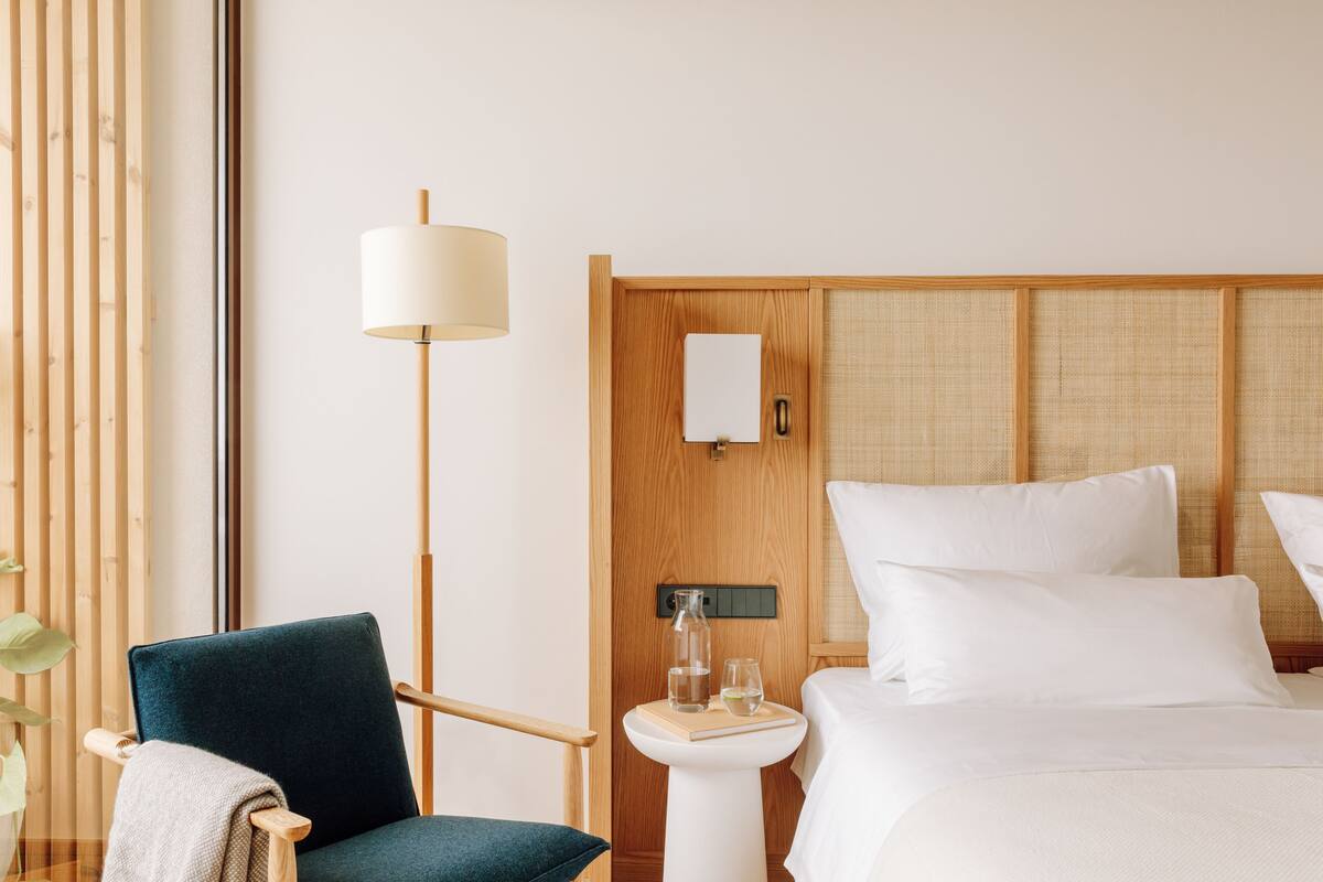 Ein Hotelzimmer mit einem Bett, einem Sessel, einem Nachttisch mit Lampe und einer an der Wand montierten Bedieneinheit.