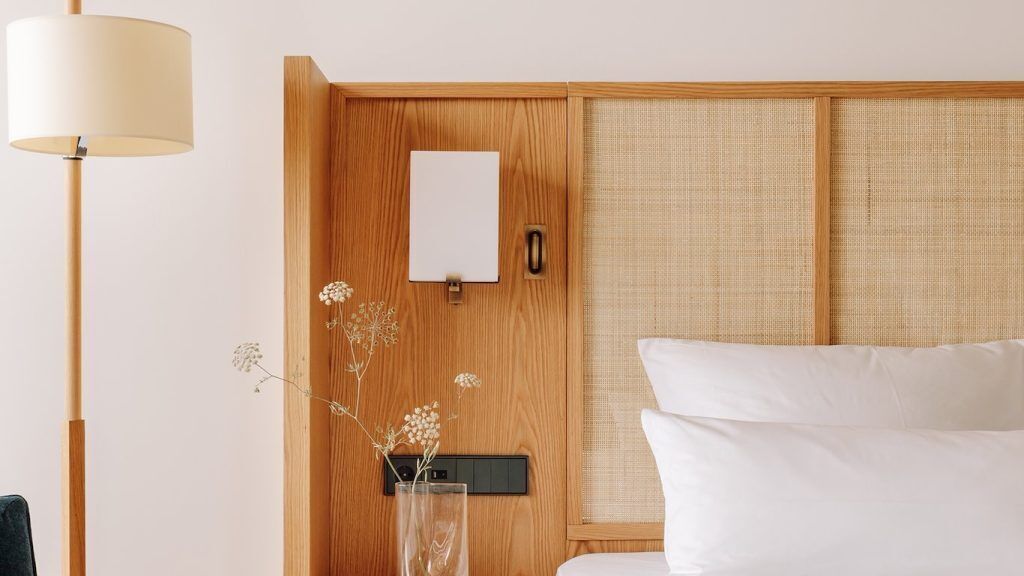 Habitación de hotel con cabecera de madera, mesita de noche con lámpara, espejo y un jarrón con flores secas.