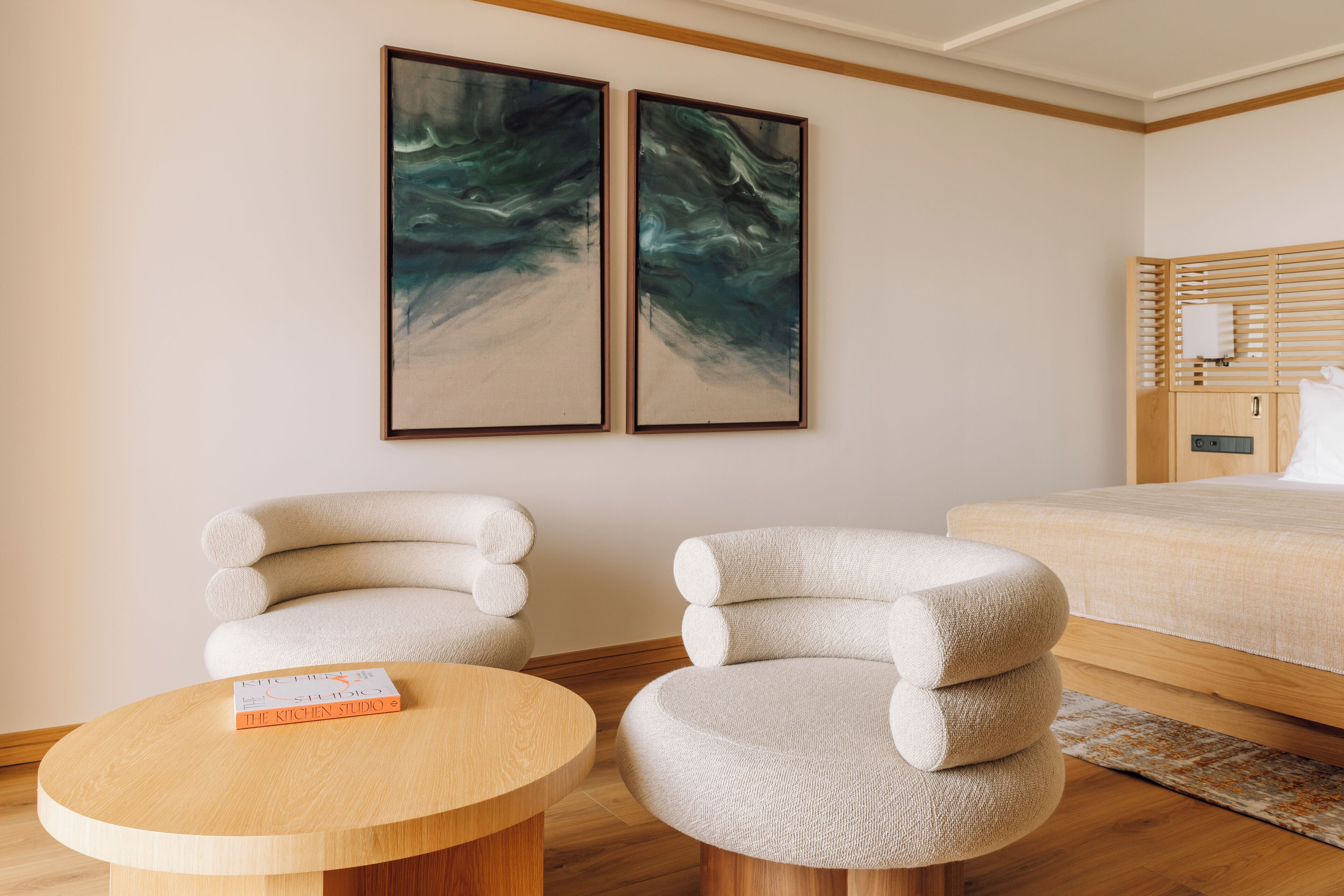 Ein modernes Hotelzimmer mit einem Bett, zwei Stühlen, einem runden Holztisch und abstrakter Wandkunst.