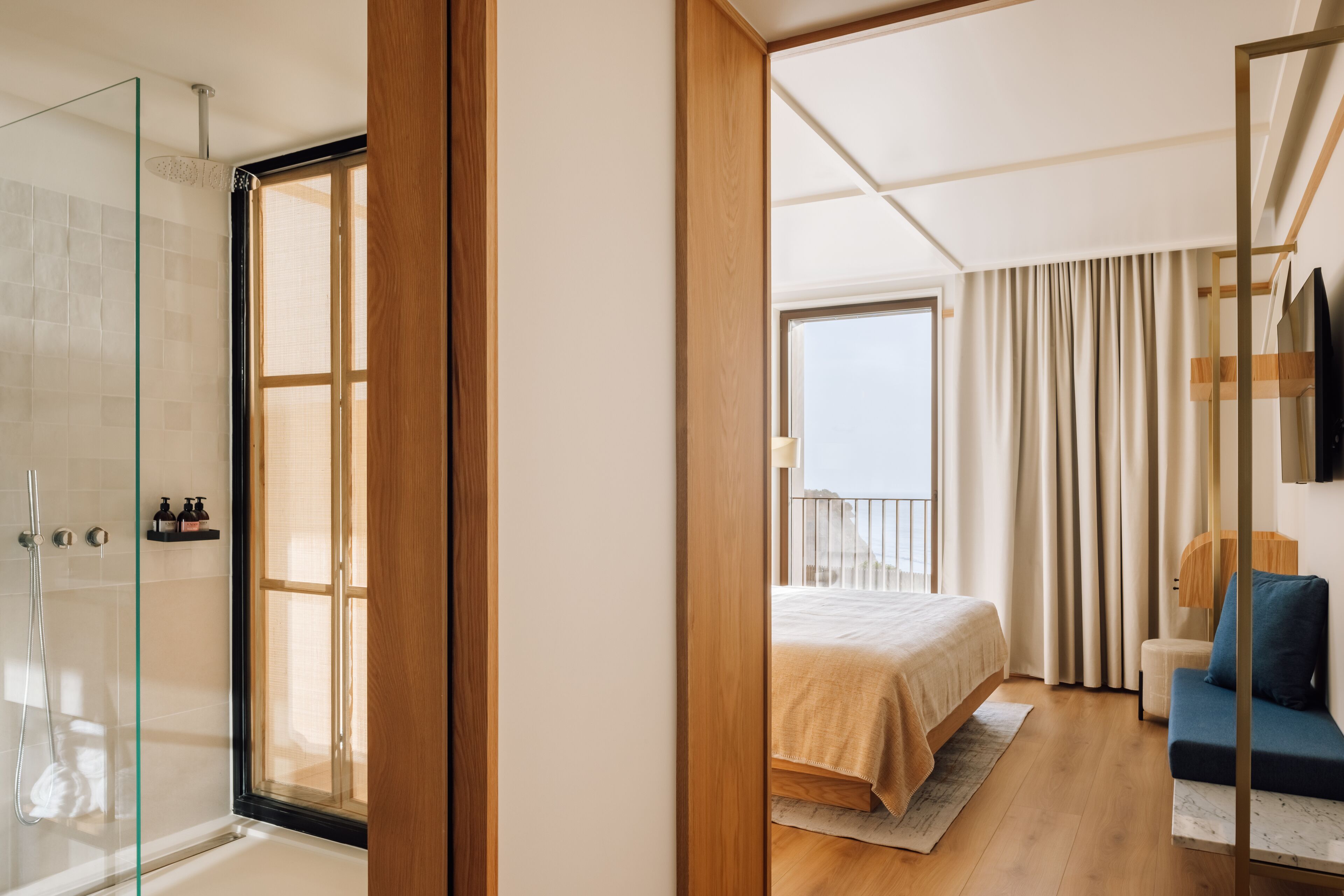A modern hotel room with a glass shower enclosure, a bed with a beige bedspread, a blue sofa, and a view of the outdoors.