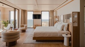 Un dormitorio moderno con una cama grande, una zona de estar con una silla y reposapiés, una mesita pequeña y un televisor montado en la pared.