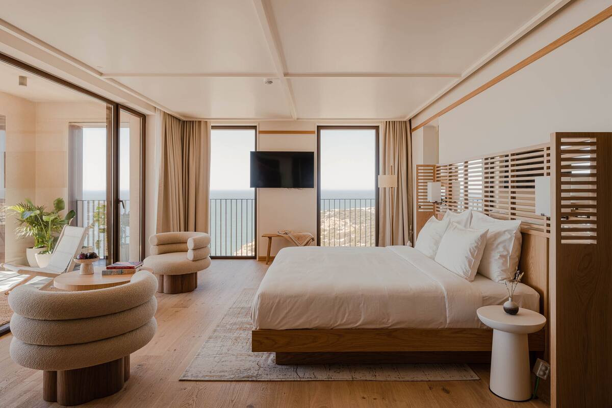 Ein modernes Schlafzimmer mit einem großen Bett, einer Sitzecke mit Sessel und Hocker, einem kleinen Tisch und einem an der Wand befestigten Fernseher.