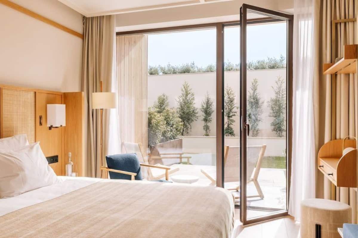 Ein Hotelzimmer mit einem großen Bett, einem Sessel, einem Tisch und einer Schiebetür zu einem Außenbereich.