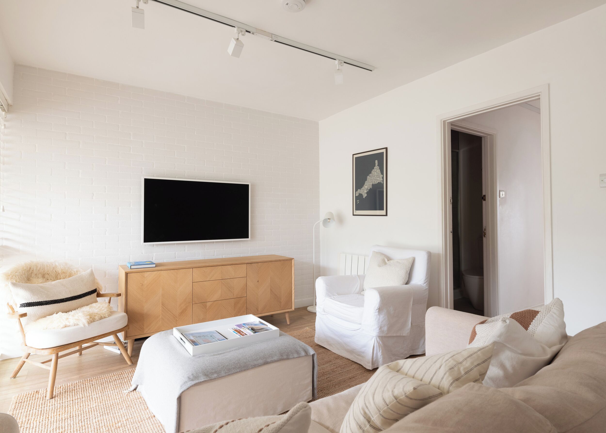 A modern living room with a white brick wall, a flat-screen TV, a wooden cabinet, a white armchair, a sofa, and a reading area with a magazine.