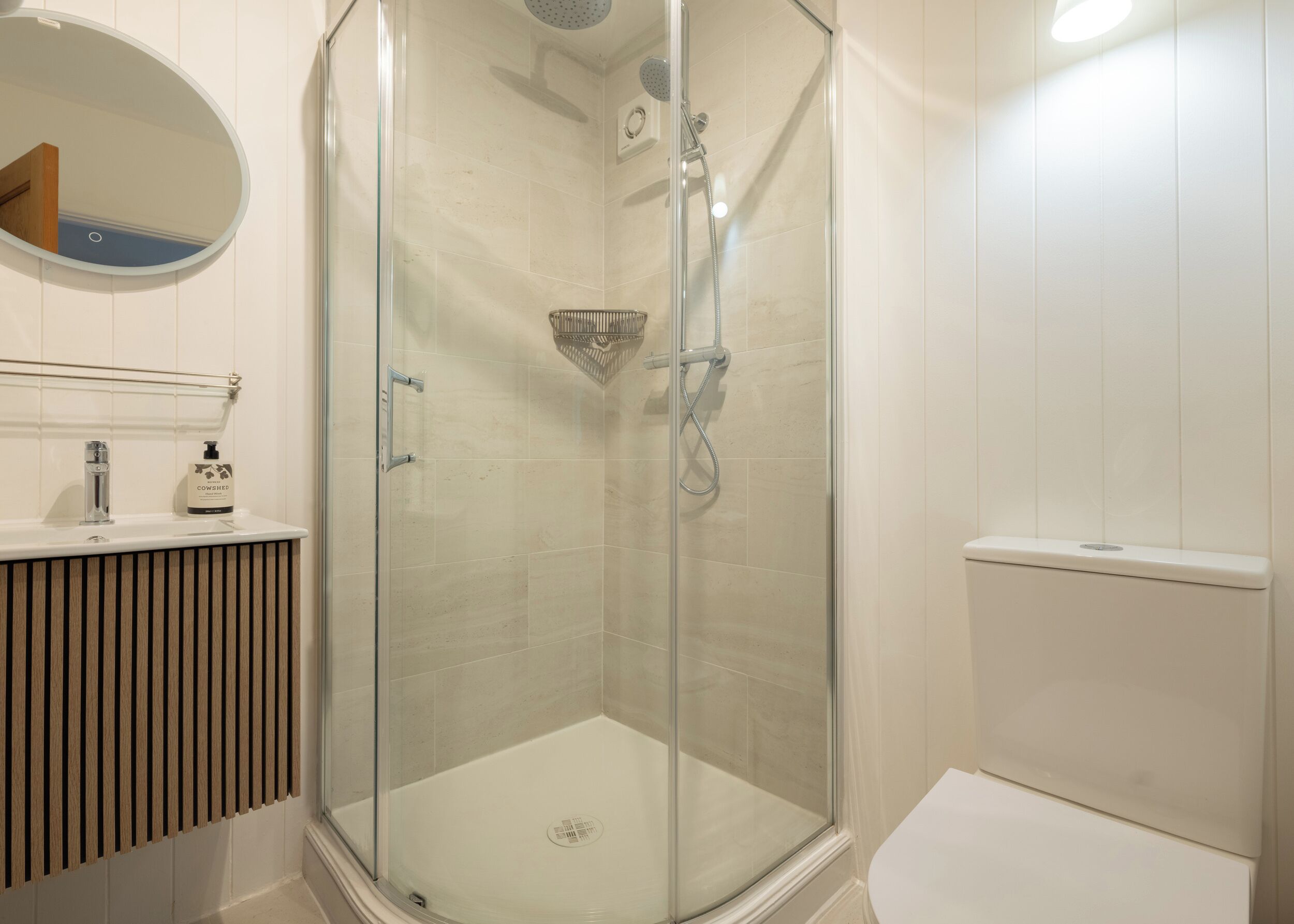 Et moderne badeværelse med en glasafskærmet brusekabine, toilet og håndvask med sæbedispenser.