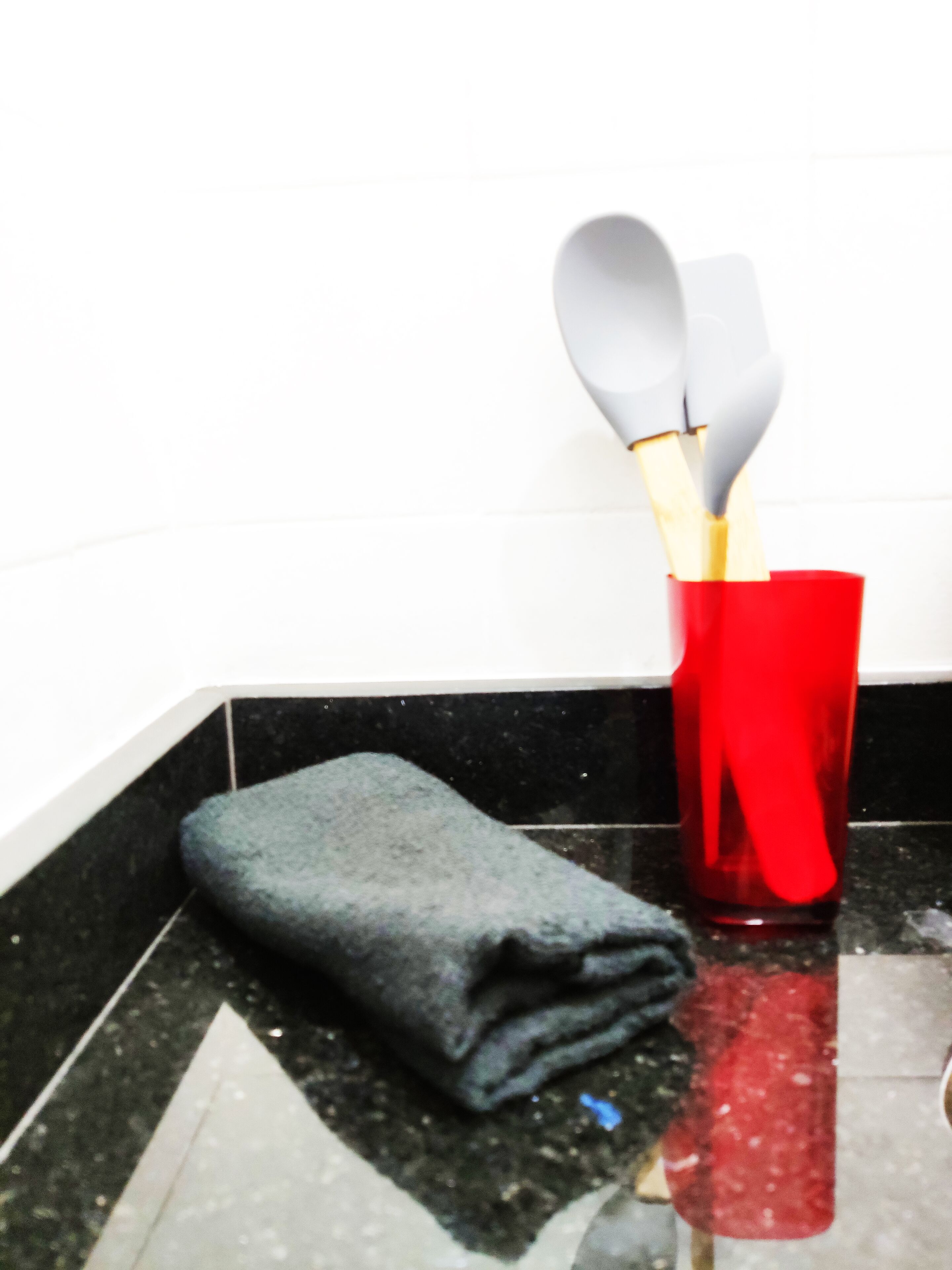 Una encimera de cocina con una taza roja que contiene una espátula y una cuchara, una toalla gris doblada y un salpicadero de baldosas blanco y negro.