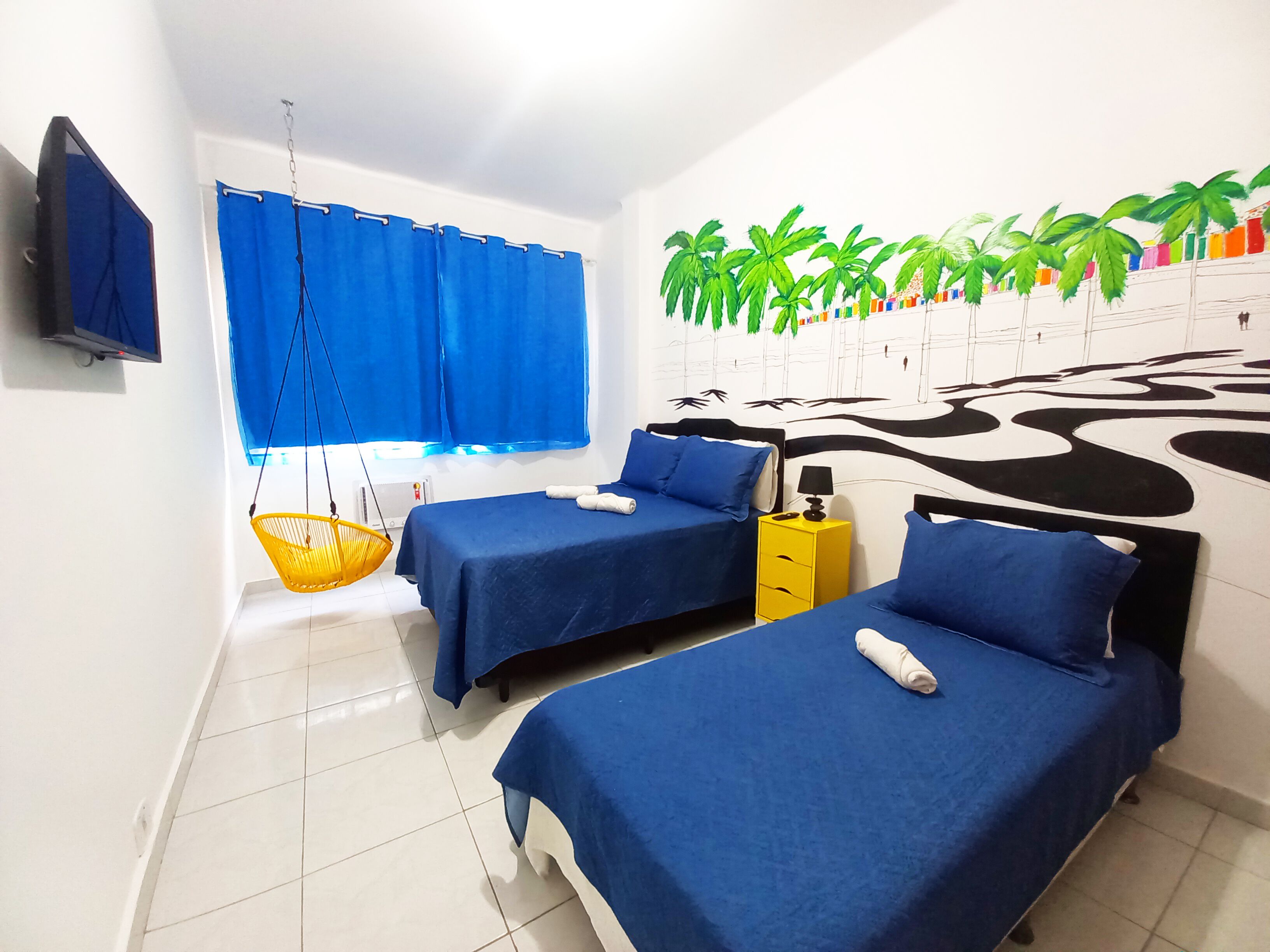 Un dormitorio con dos camas, televisor, una silla colgante y un mural en la pared.
