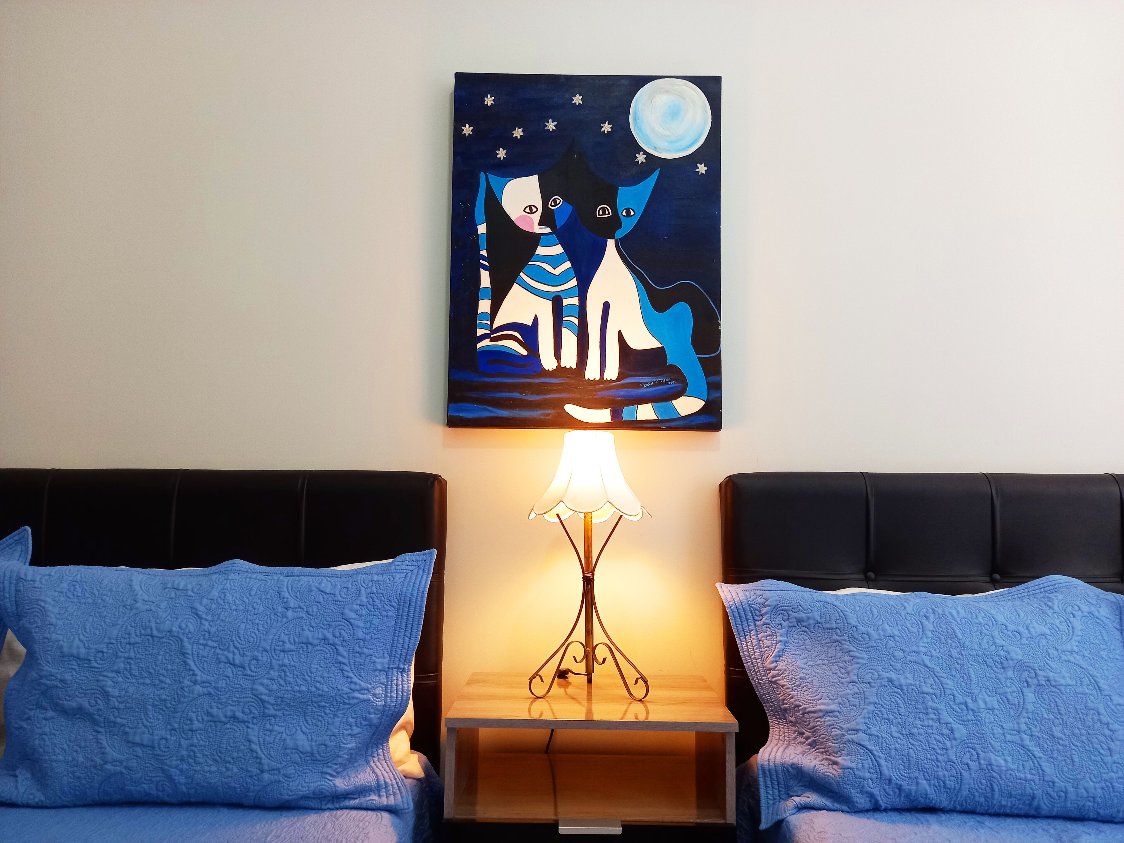 Una camera da letto con un letto, due cuscini azzurri, un comodino con una lampada e un quadro appeso al muro raffigurante una scena illuminata dalla luna.