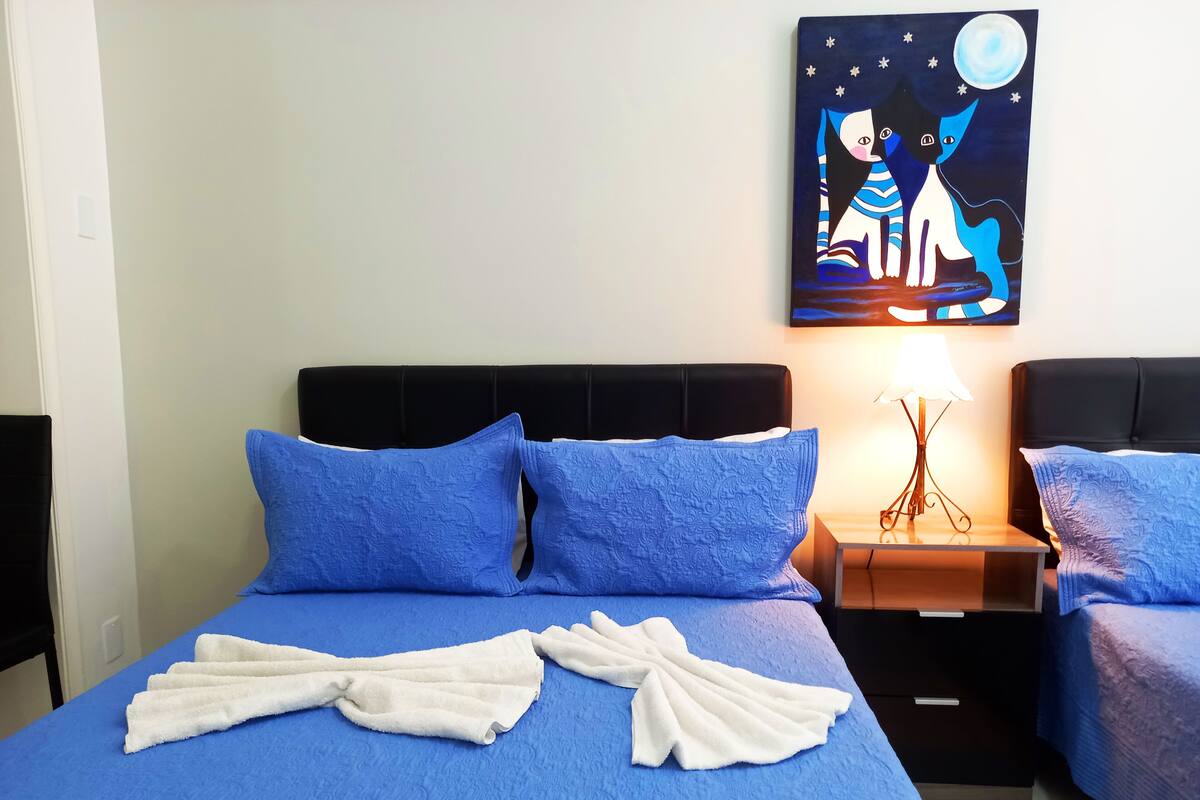 Habitación de hotel con cama, dos almohadas, mesita de noche con lámpara y un cuadro en la pared.