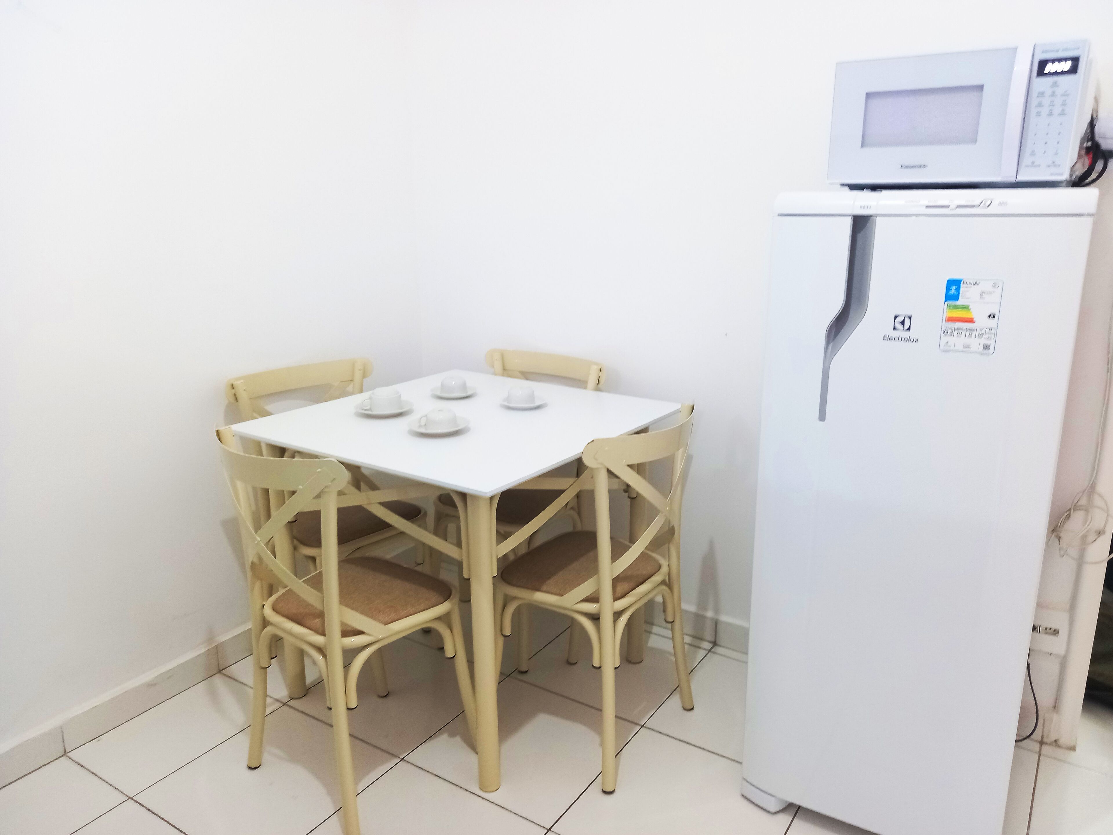 A small kitchenette with a white refrigerator and microwave, a white table with four white cups, and four beige chairs.