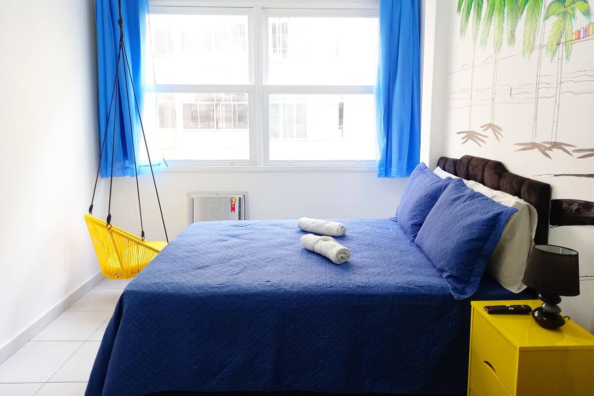 Un dormitorio con una cama, una mesita de noche amarilla, una silla colgante y una ventana con cortinas azules.