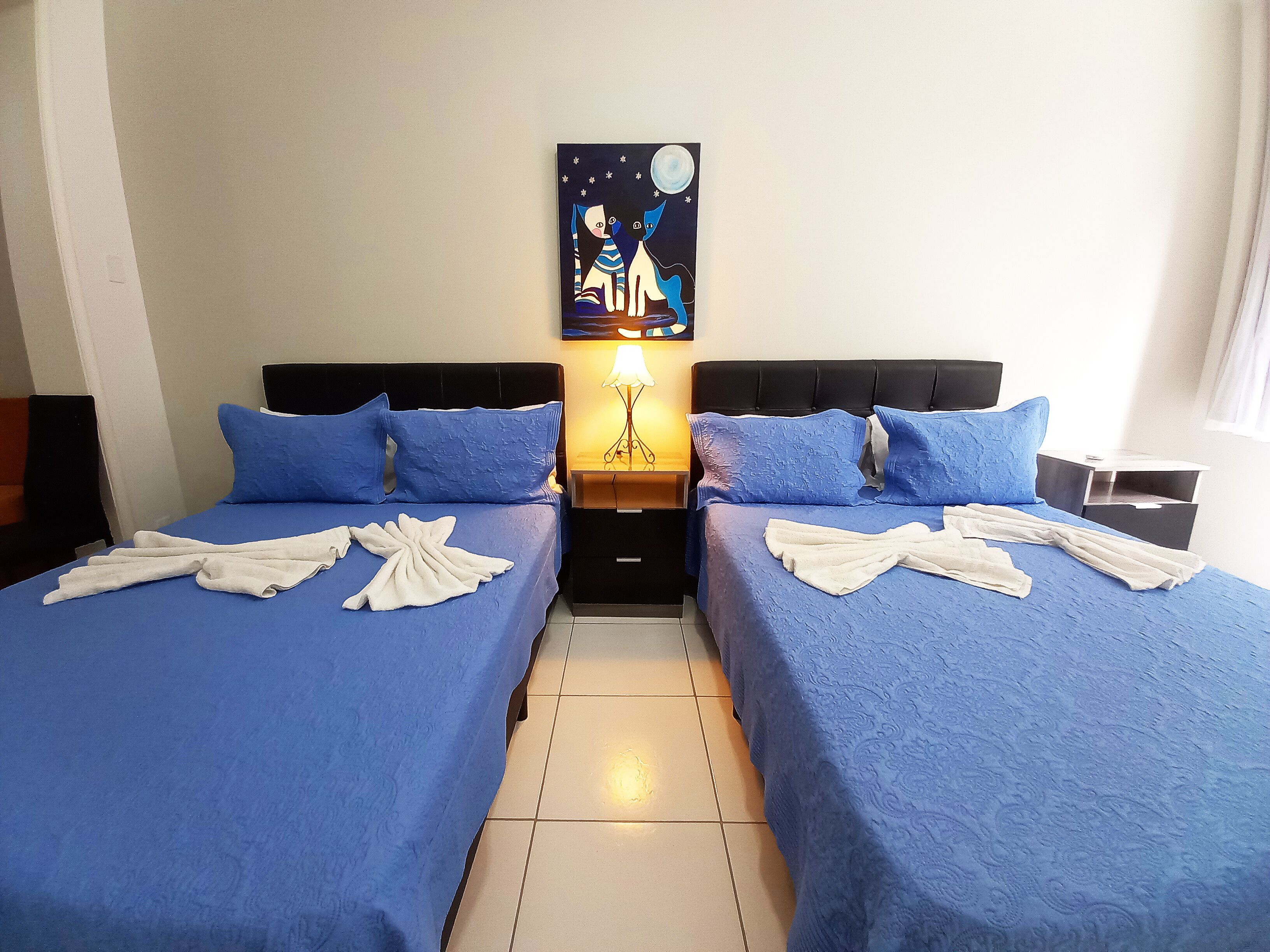 Een hotelkamer met twee bedden, elk voorzien van blauw beddengoed en witte handdoeken. Op de muur hangt een schilderij van een kat en een maan.