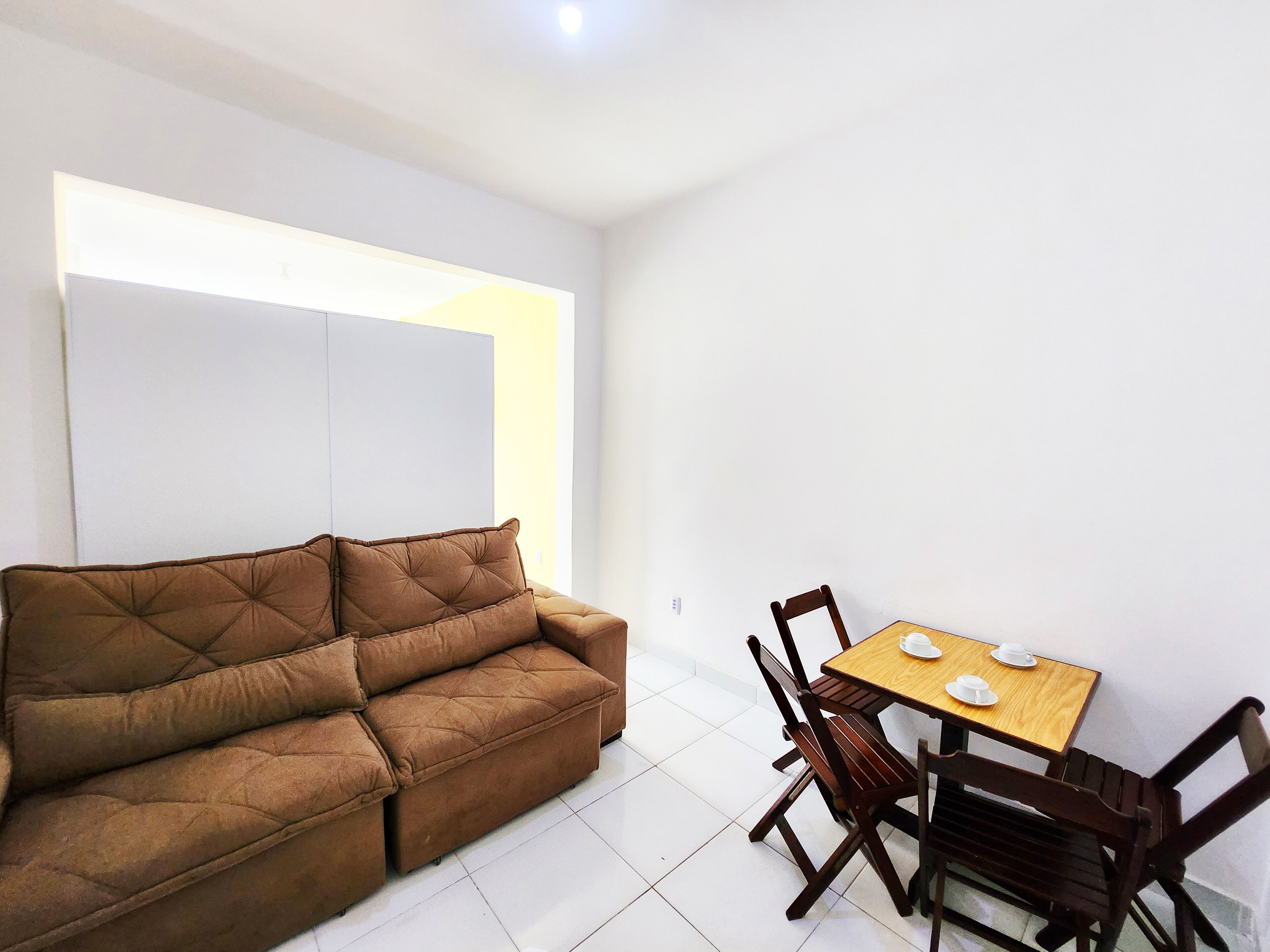 A living room with a brown sofa, a wooden dining table with four chairs, and a white wall.