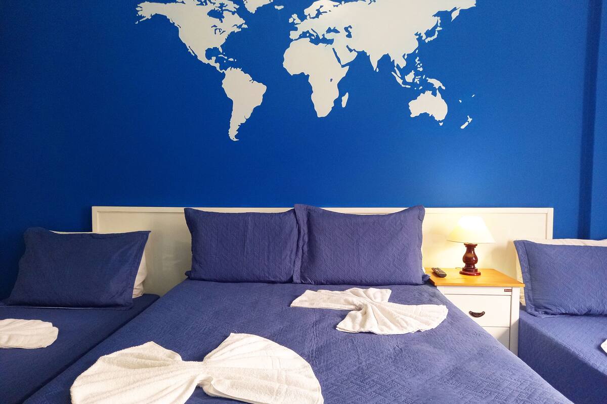 Habitación de hotel con un mapa del mundo en la pared, dos camas con ropa de cama azul y una mesita de noche con una lámpara.