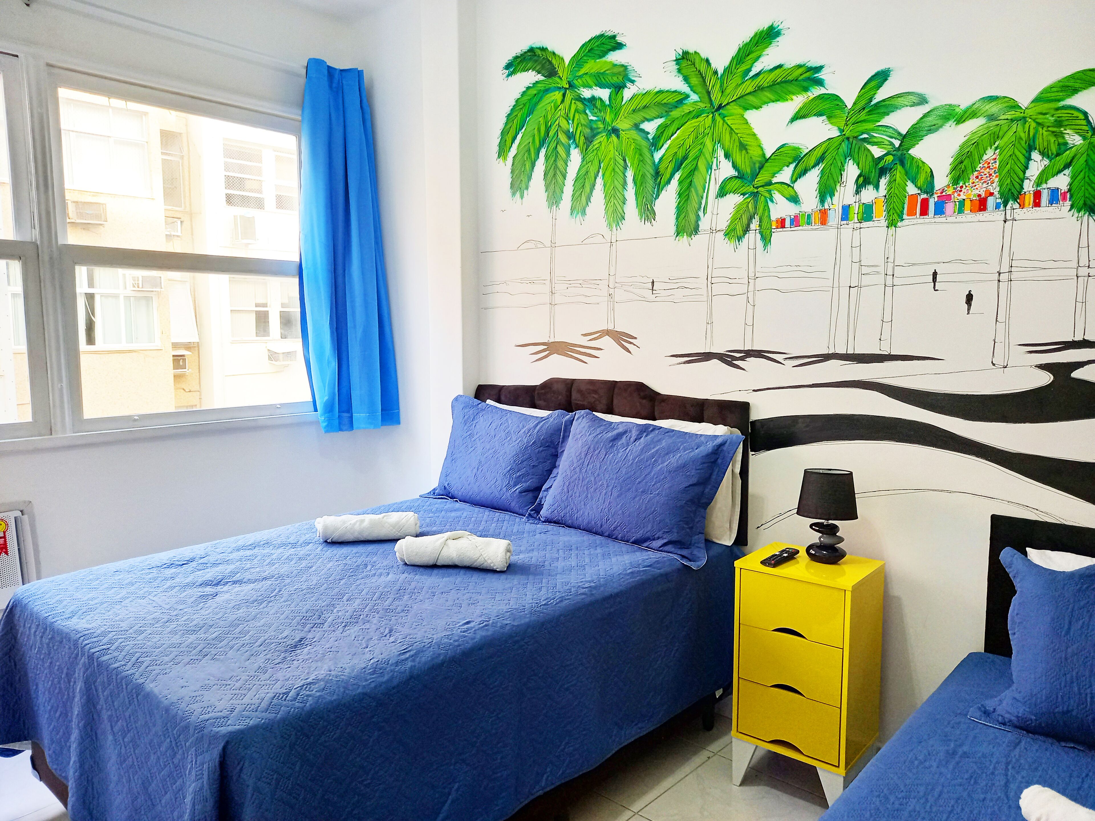 A bedroom with a bed, a nightstand, a wall mural, and a window with curtains.