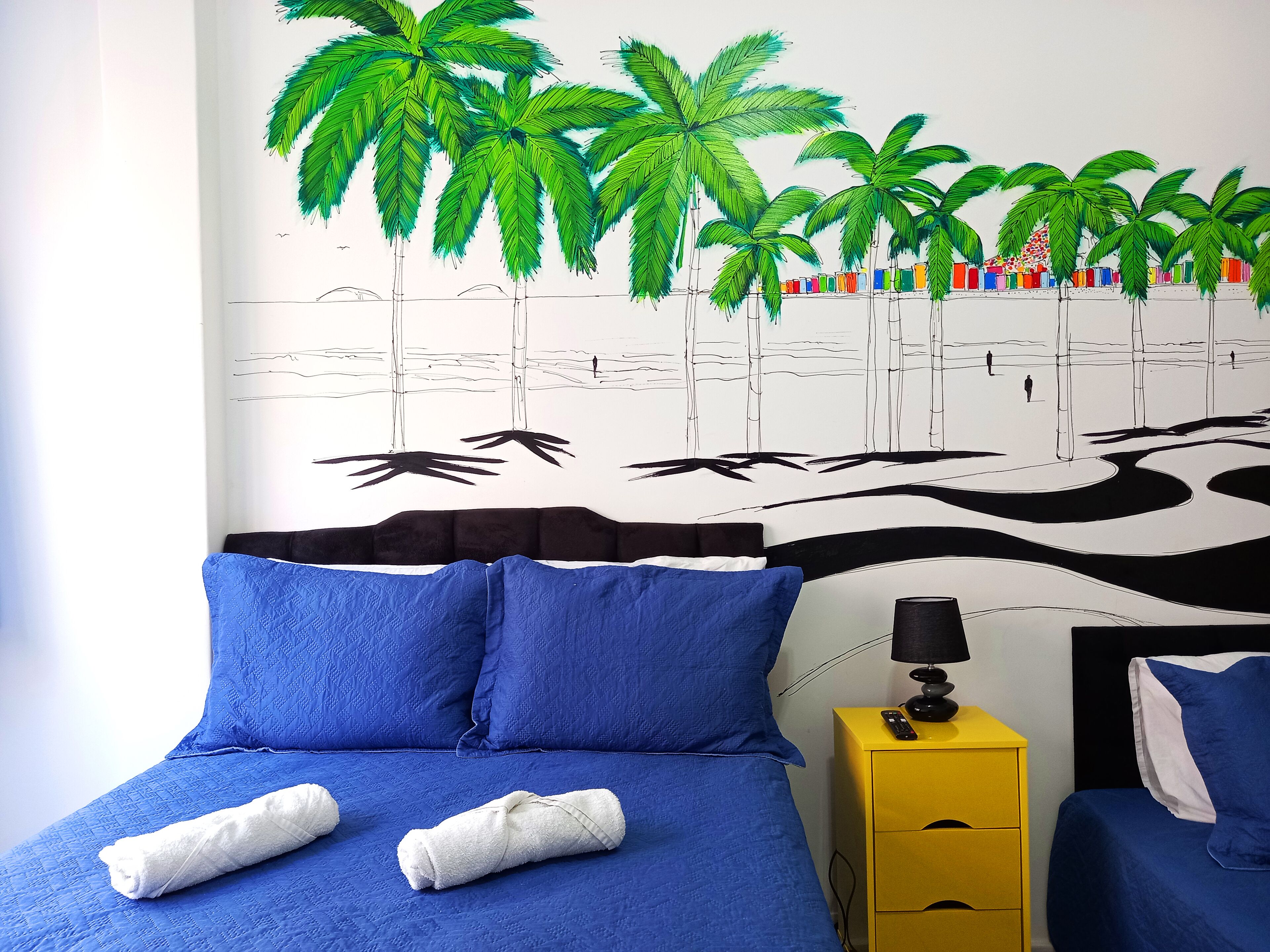 Ein Schlafzimmer mit einem Bett, blauen Kissen, einem gelben Nachttisch und einer Wandmalerei von Palmen.