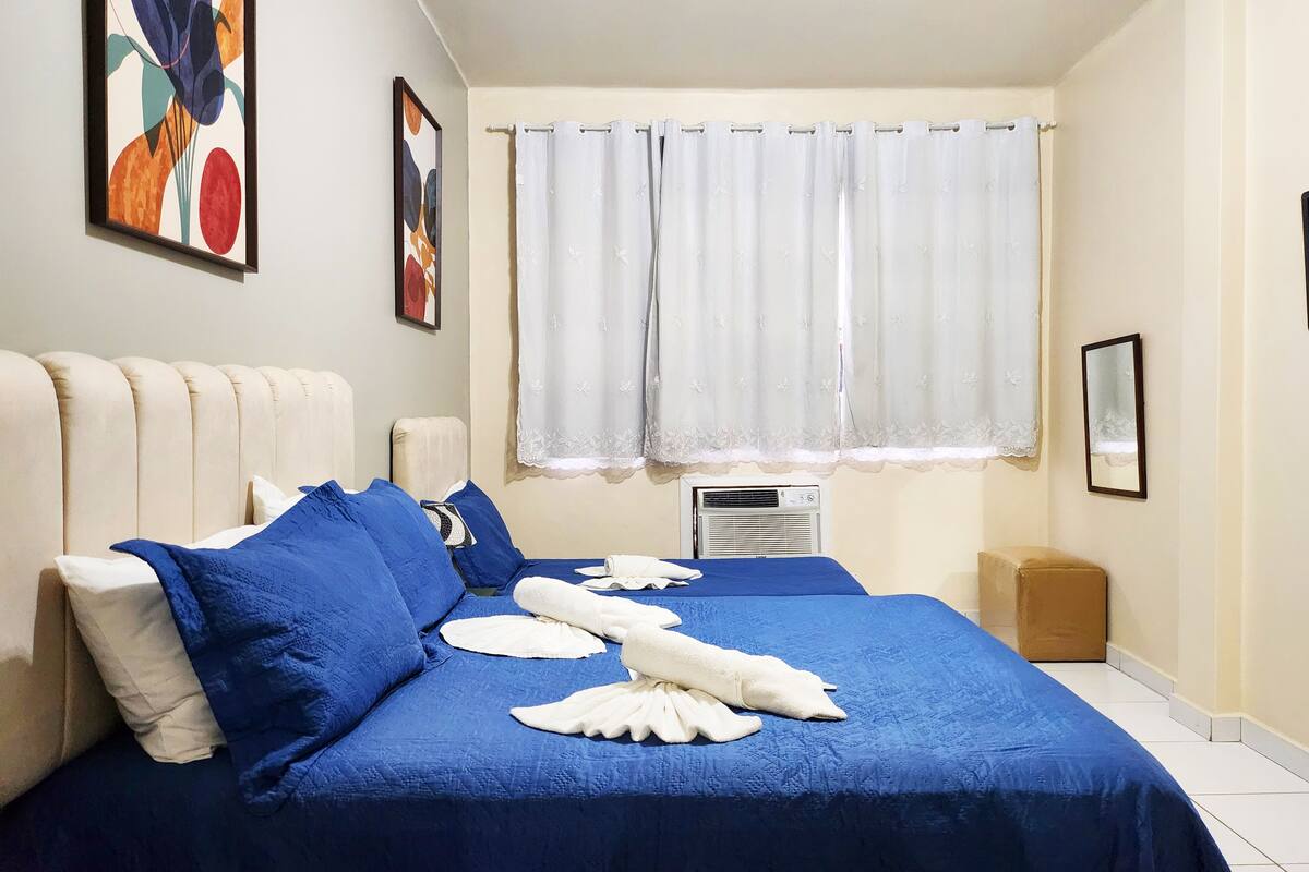 Habitación de hotel con cama, almohadas, ventana con cortinas, aire acondicionado de pared y un cuadro enmarcado.