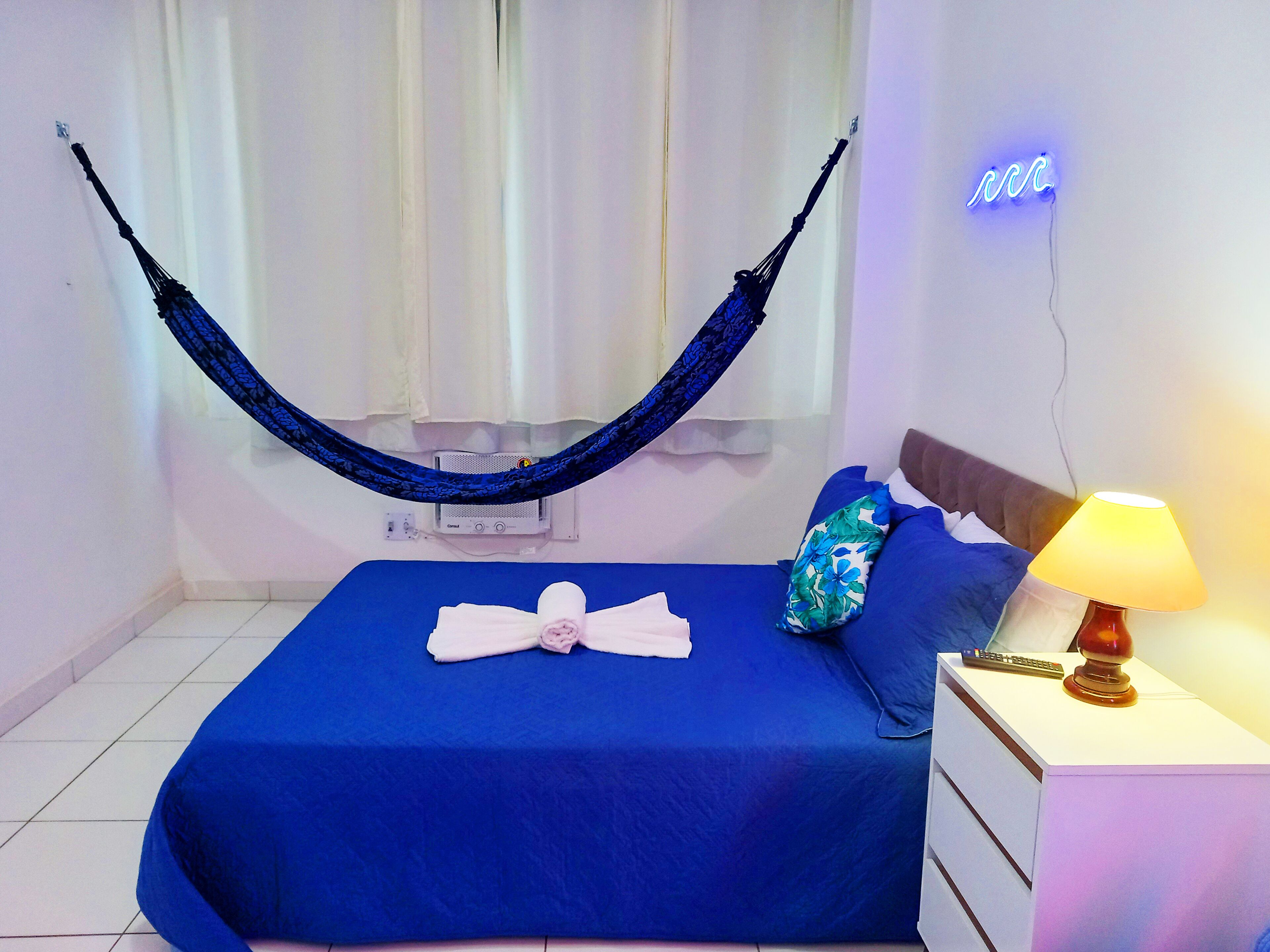 A neatly made bed with a blue bedspread, a hammock, and a bedside lamp.