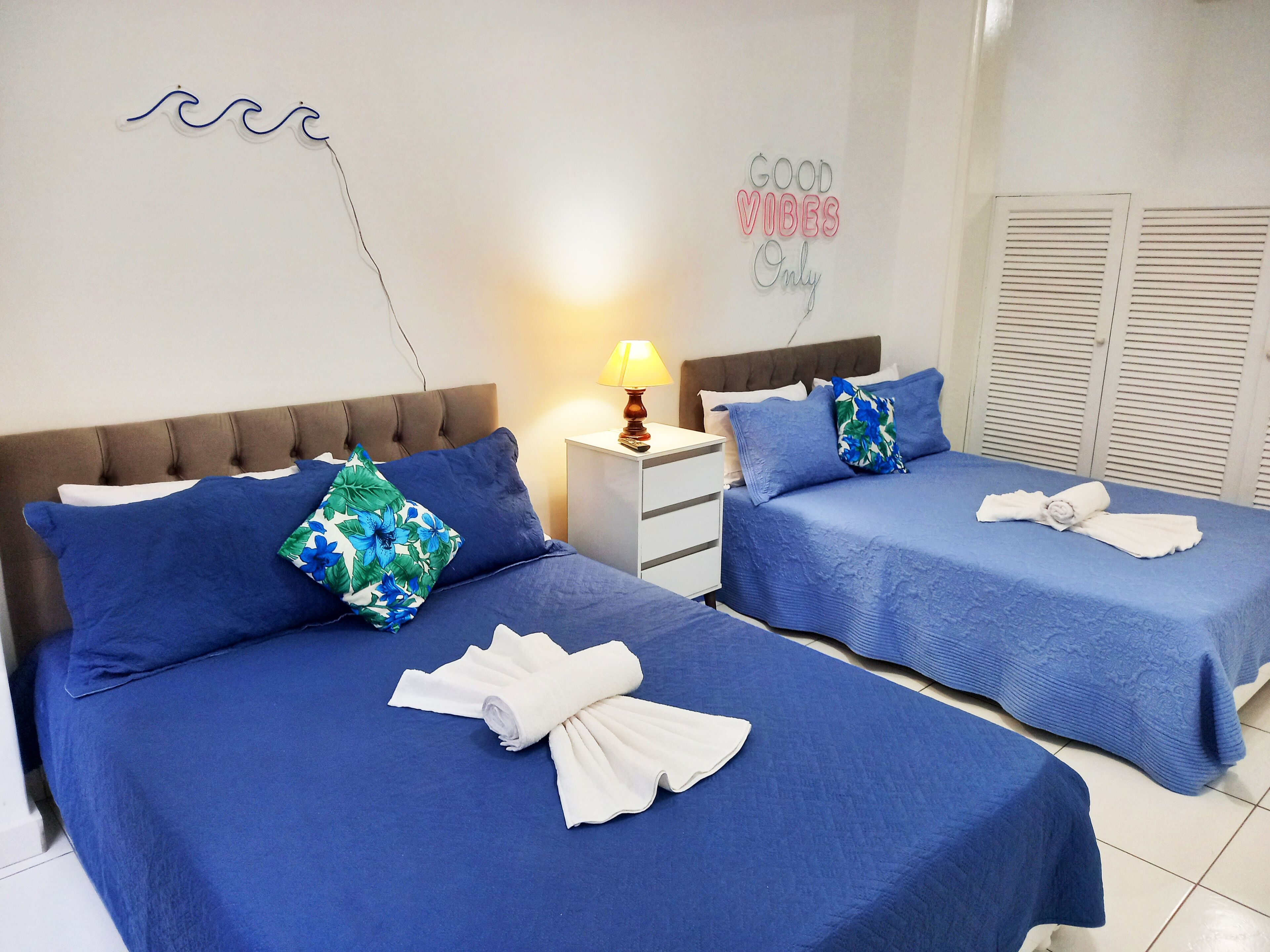A hotel room with two beds, each with blue bedding and a floral pillow. There is a nightstand with a lamp, a wall decoration with the phrase "GOOD VIBES Only", and a towel on each bed.