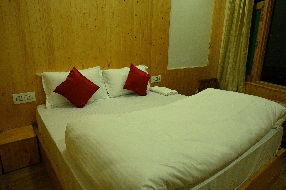 A neatly made bed with white bedding and red pillows in a room with wooden walls and a window with curtains.