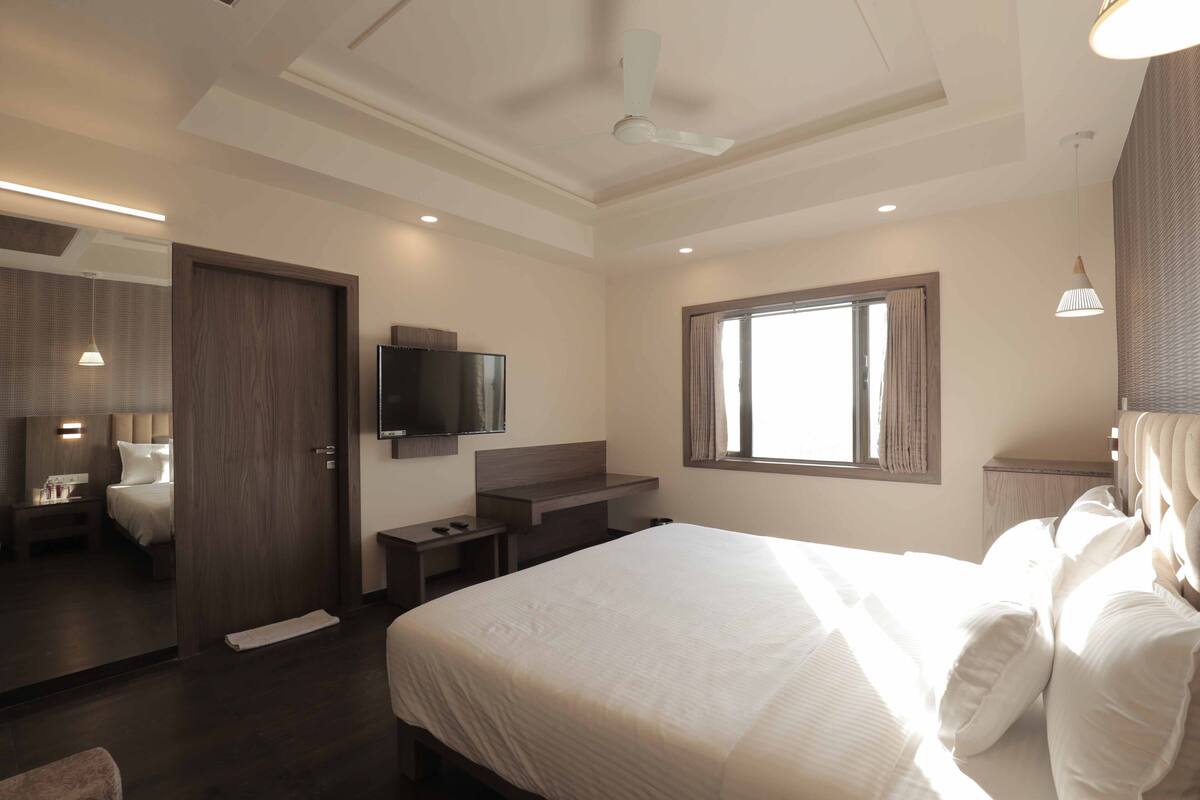 A modern hotel room with a large bed, a flat-screen TV, a window with curtains, a ceiling fan, and a small desk with a chair.