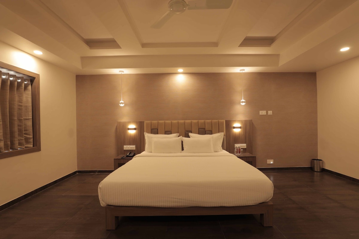 A modern hotel room with a large bed, wooden headboard, recessed ceiling lights, and a small bedside table with a lamp.
