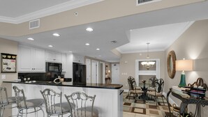 A modern kitchen with white cabinets, a black countertop, and stainless steel appliances. There are three bar stools and a dining table with chairs in the adjacent dining area. - 2311-23rd Flr-Outdoor Pool-Poolside Cafe-Hot Tub-Grills-AMAZING Birds Eye VIEW! (Panama City Beach)