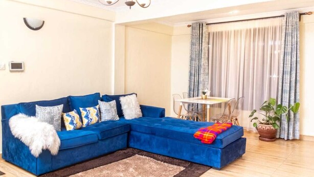 A living room with a blue sectional sofa, a small dining table, and a potted plant. - Stay.Plus Ruaka View Apartment (Ruaka)