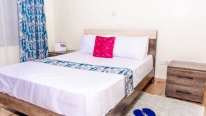 A neatly made bed with a white and blue patterned bedspread, a red pillow, and a wooden nightstand with a drawer. - Stay.Plus Ruaka View Apartment (Ruaka)