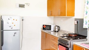 A compact kitchen with wooden cabinets, a white refrigerator, a microwave, a gas stove, and a kettle. - Stay.Plus Ruaka View Apartment (Ruaka)