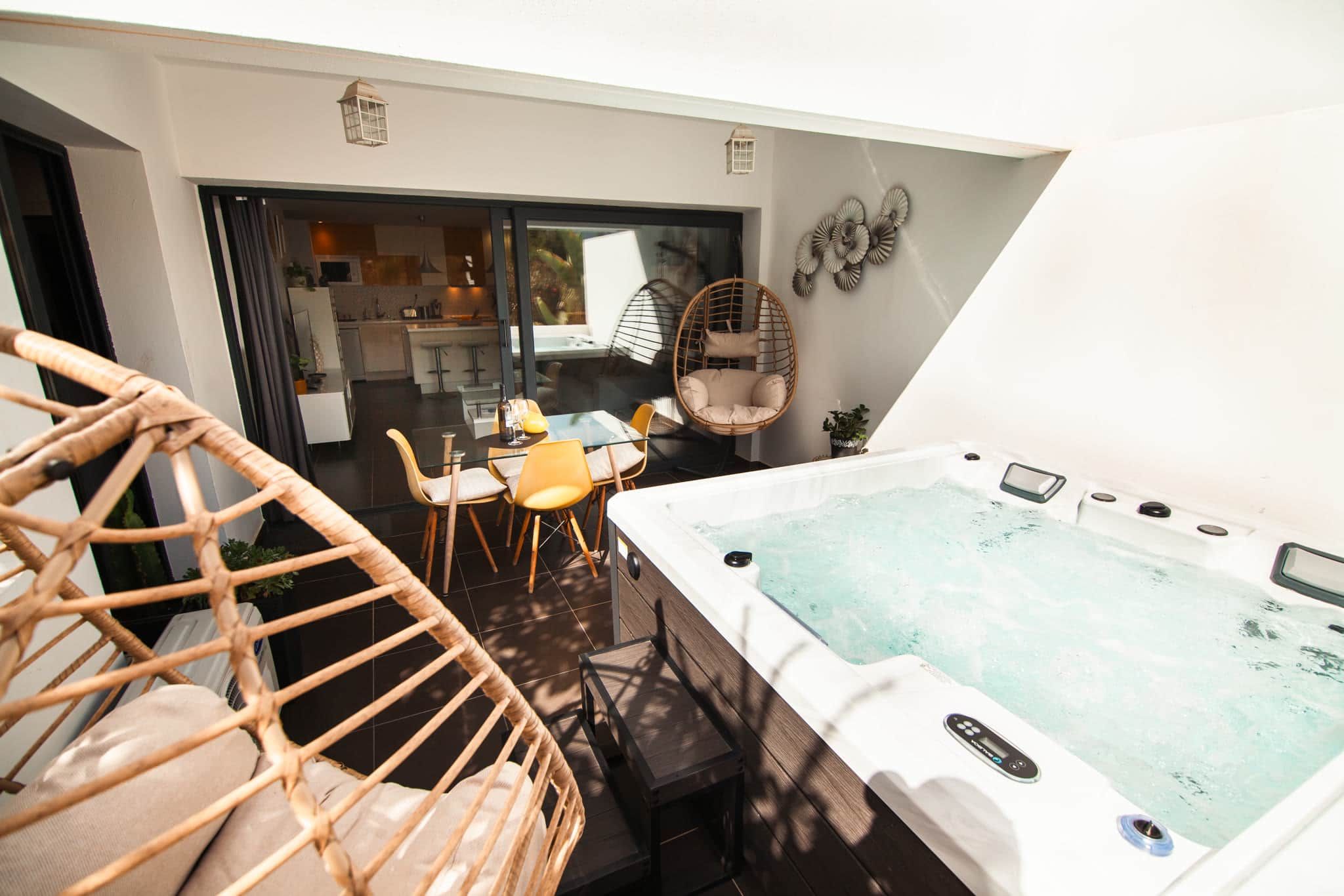 Eine moderne Außenpatio mit Whirlpool, Korbstühlen, einem Esstisch und einem durch Glastüren sichtbaren Küchenbereich.