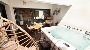 A modern outdoor patio with a hot tub, wicker chairs, a dining table, and a kitchen area visible through glass doors.