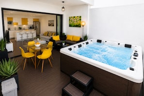 Un espacio exterior moderno con jacuzzi, sillas amarillas, una mesa de vidrio y un sofá con cojines amarillos.