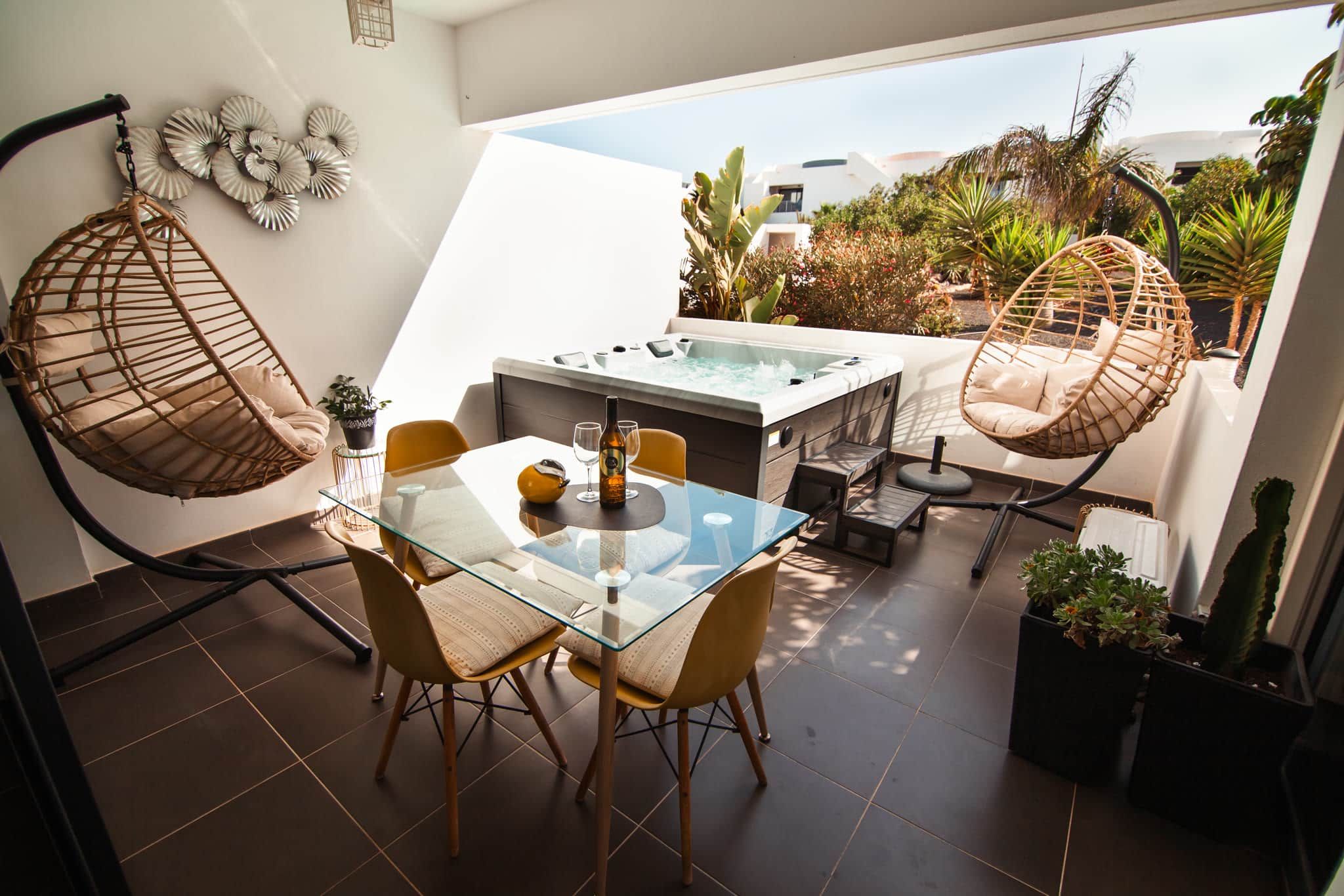 Une terrasse extérieure moderne avec une table en verre, deux fauteuils en osier, un jacuzzi et des plantes en pot.