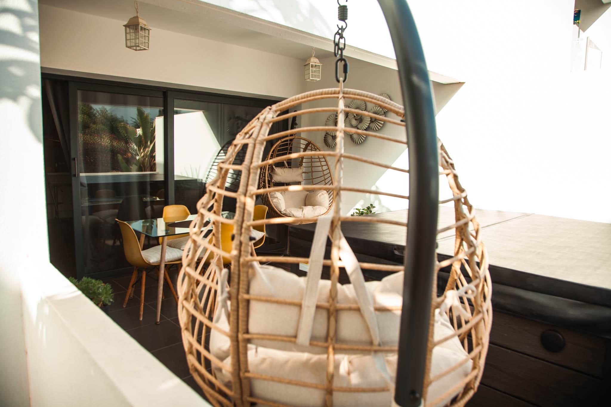 A wicker hanging chair with cushions, a glass table, and chairs in a modern outdoor setting.