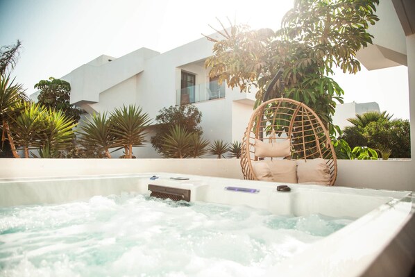 A hot tub with a wicker chair and cushions, surrounded by tropical plants and a modern building in the background.