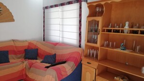 A room with a striped sofa, a wooden cabinet with glassware, and a window with a floral curtain. - Les Motes 2º Solo Familias! (Xeraco Playa)