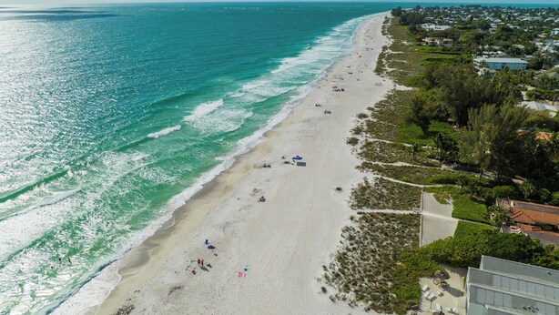 An aerial view of a beach with clear turquoise waters, a sandy shore, and a row of buildings along the coastline.