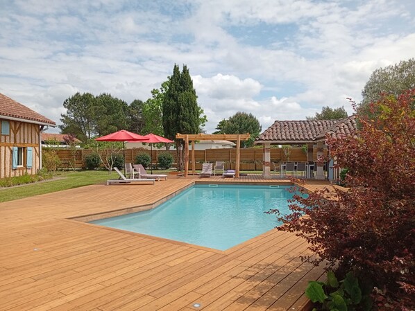 Une terrasse en bois avec une piscine, des chaises longues et une pergola surmontée de parasols rouges.