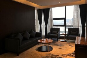 A modern hotel room with a dark-colored sofa, two armchairs, a small table, and a large window with curtains.