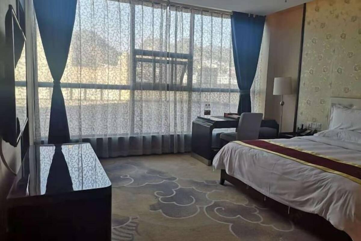 A hotel room with a bed, a desk, a chair, a lamp, and a large window with curtains.