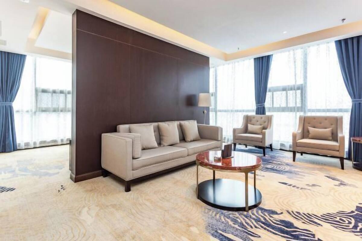 A modern hotel room with a sofa, armchairs, a coffee table, and large windows with curtains.