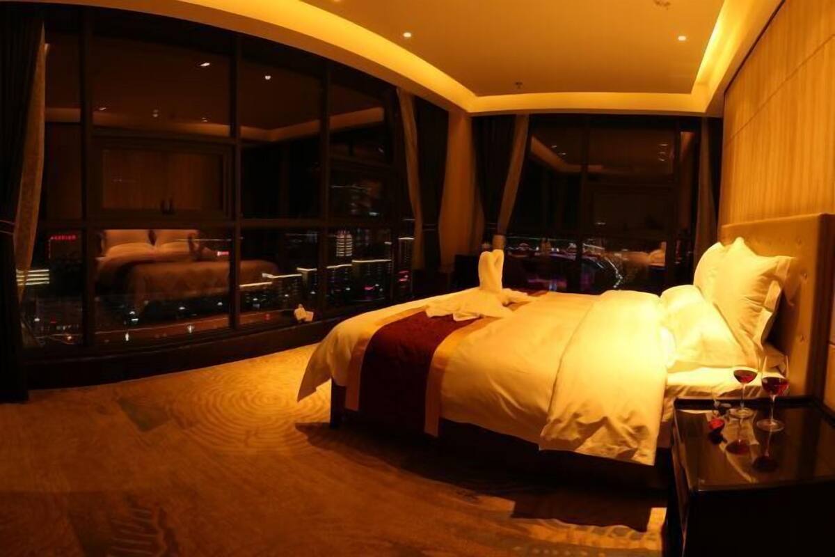 A hotel room with a large bed, a nightstand, and a view through large windows.